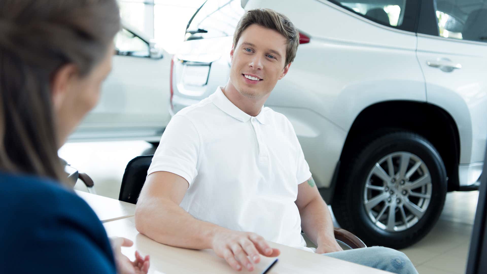 一位身穿白色马球衫的准男性买家坐在一家汽车经销商的办公桌旁。